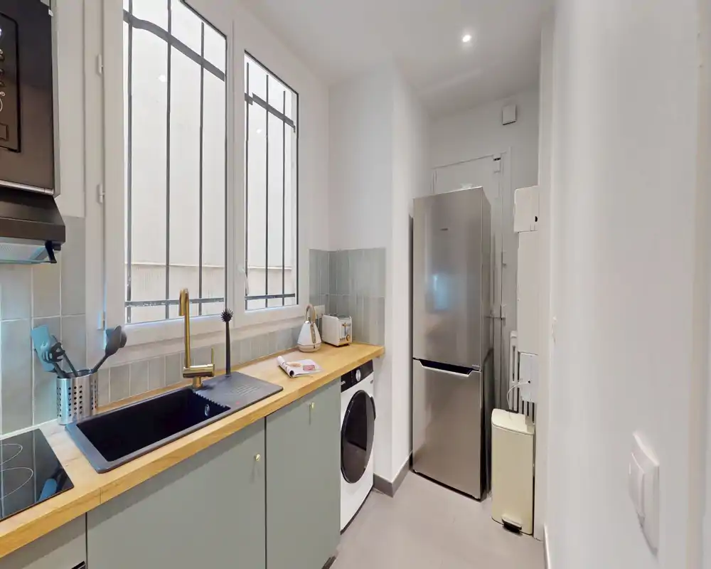 Cuisine en longueur moderne et compacte avec plan de travail en bois, évier intégré, plaque à induction et réfrigérateur haut ; lumière naturelle qui la rend accueillante.