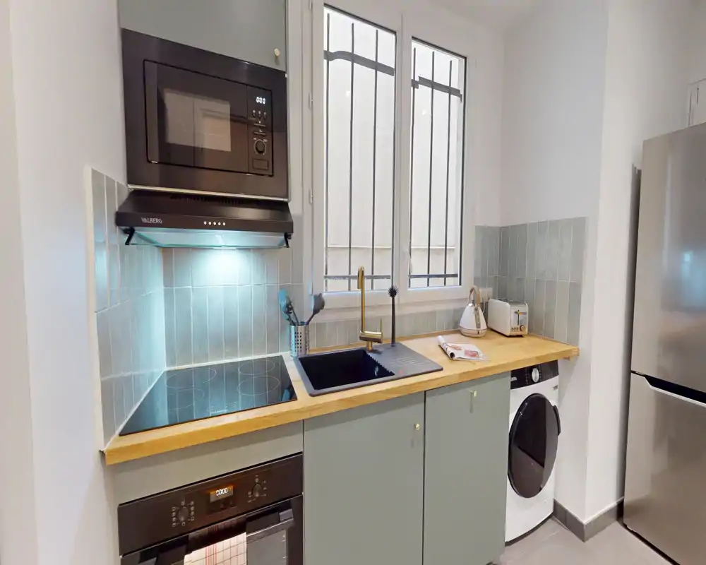 Cuisine compacte et moderne avec plan de travail en bois, plaque à induction intégrée, évier sous la fenêtre, micro-ondes et électroménagers. Espace propre et lumineux, parfait pour les locations courtes.