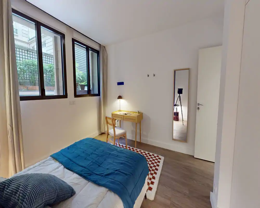 Chambre moderne et lumineuse avec un lit double bien fait, une couette bleue, un petit bureau en bois avec lampe et une glace pleine longueur — bien éclairée par de grandes fenêtres donnant sur une cour végétalisée.