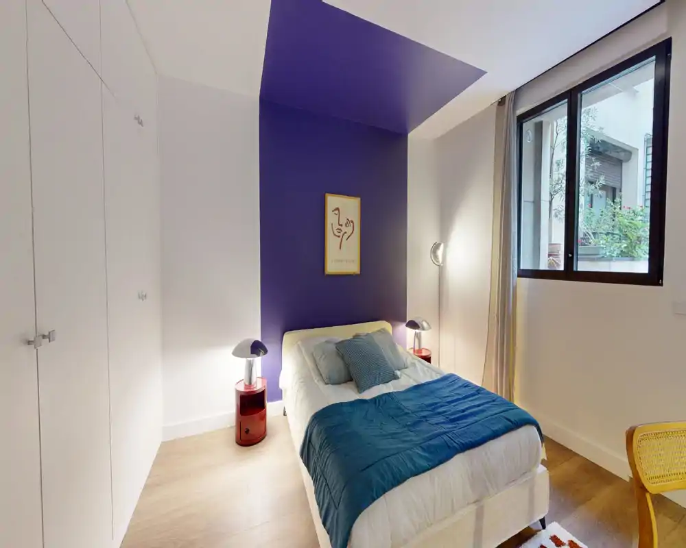 Chambre moderne et lumineuse avec un lit simple central contre un mur violet accentué, lampes de chevet, placard intégré et fenêtre laissant entrer la lumière naturelle.