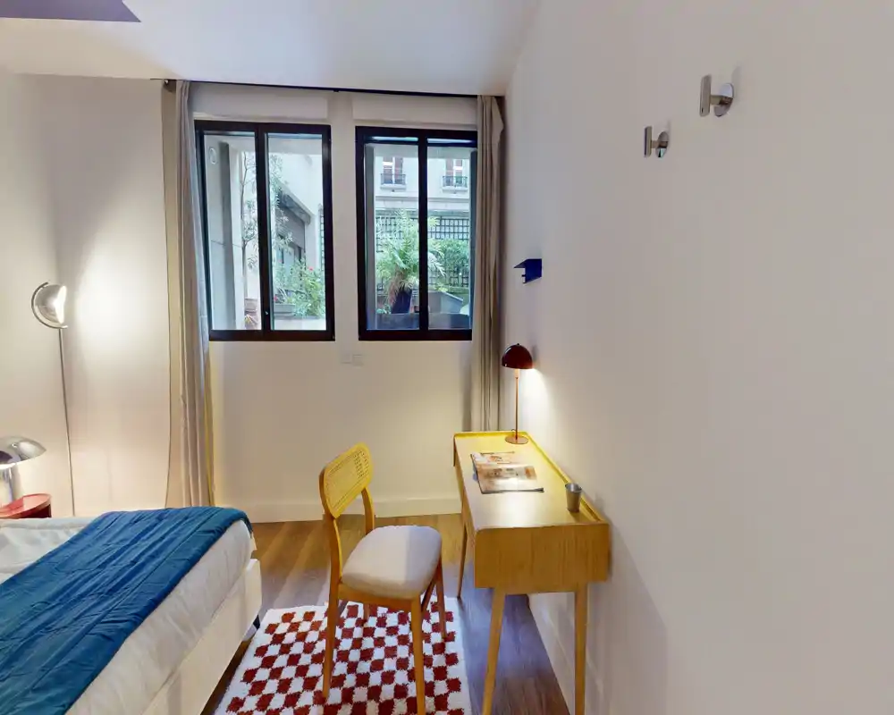 Chambre moderne et lumineuse avec lit visible, bureau en bois et chaise près de la fenêtre ; textiles chaleureux et lampes créent une ambiance accueillante.