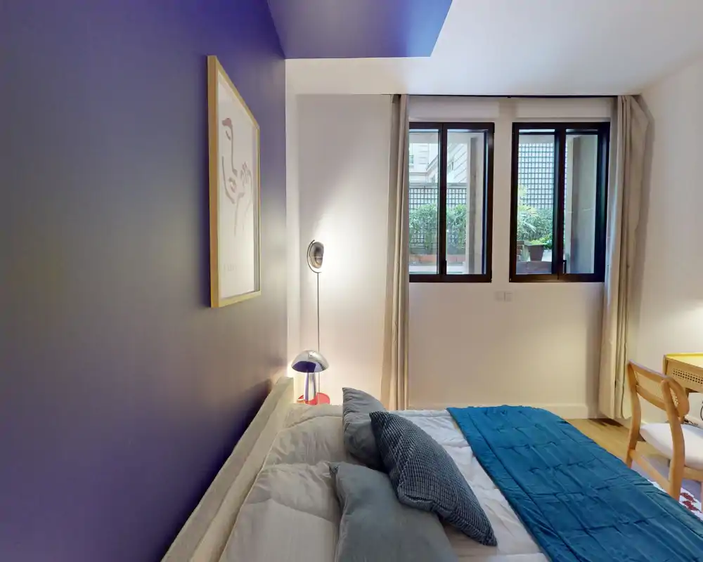 Chambre moderne et lumineuse avec un mur d’accent violet, lit double habillé de coussins et d’un plaid turquoise. La lumière naturelle des fenêtres et les lampes de chevet apportent une atmosphère chaleureuse, idéale pour une annonce.