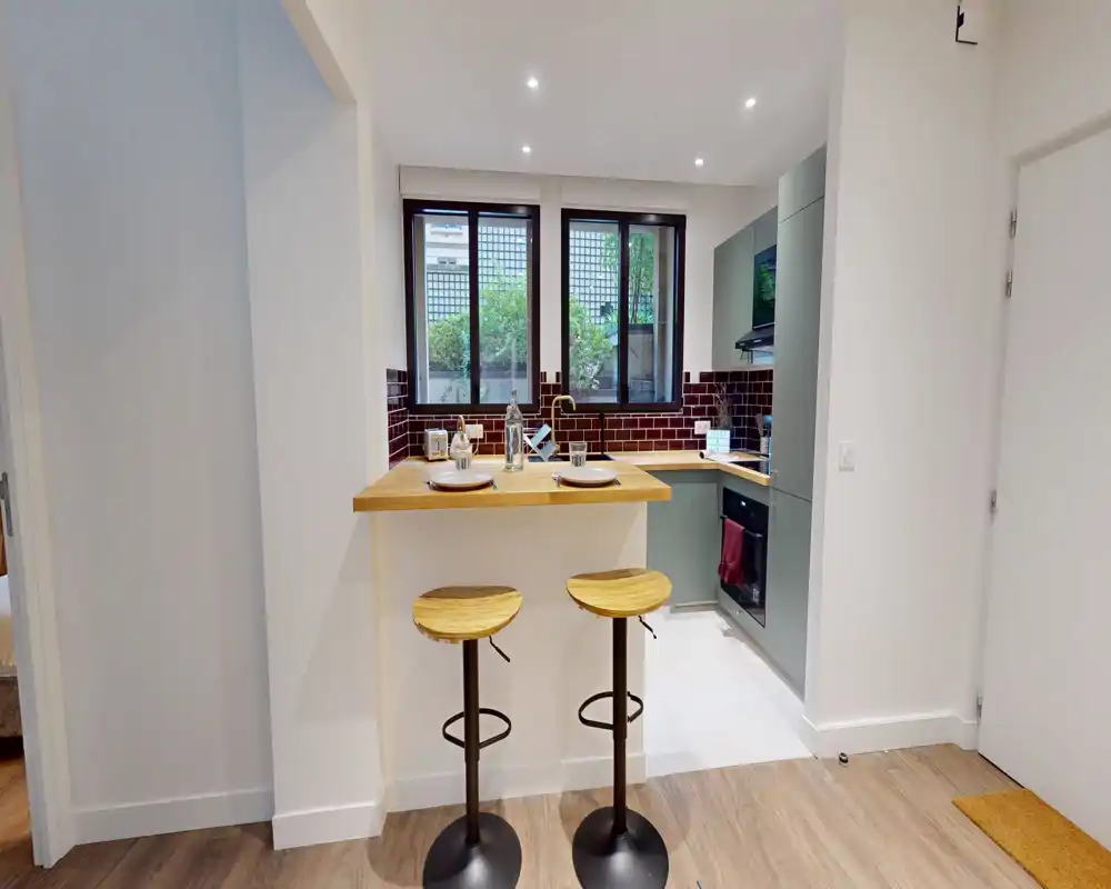 Cuisine moderne et compacte avec bar petit-déjeuner et deux tabourets en bois, placards verts et crédence en carreaux bordeaux ; espace de cuisson lumineux et bien organisé avec fenêtres donnant sur la verdure.