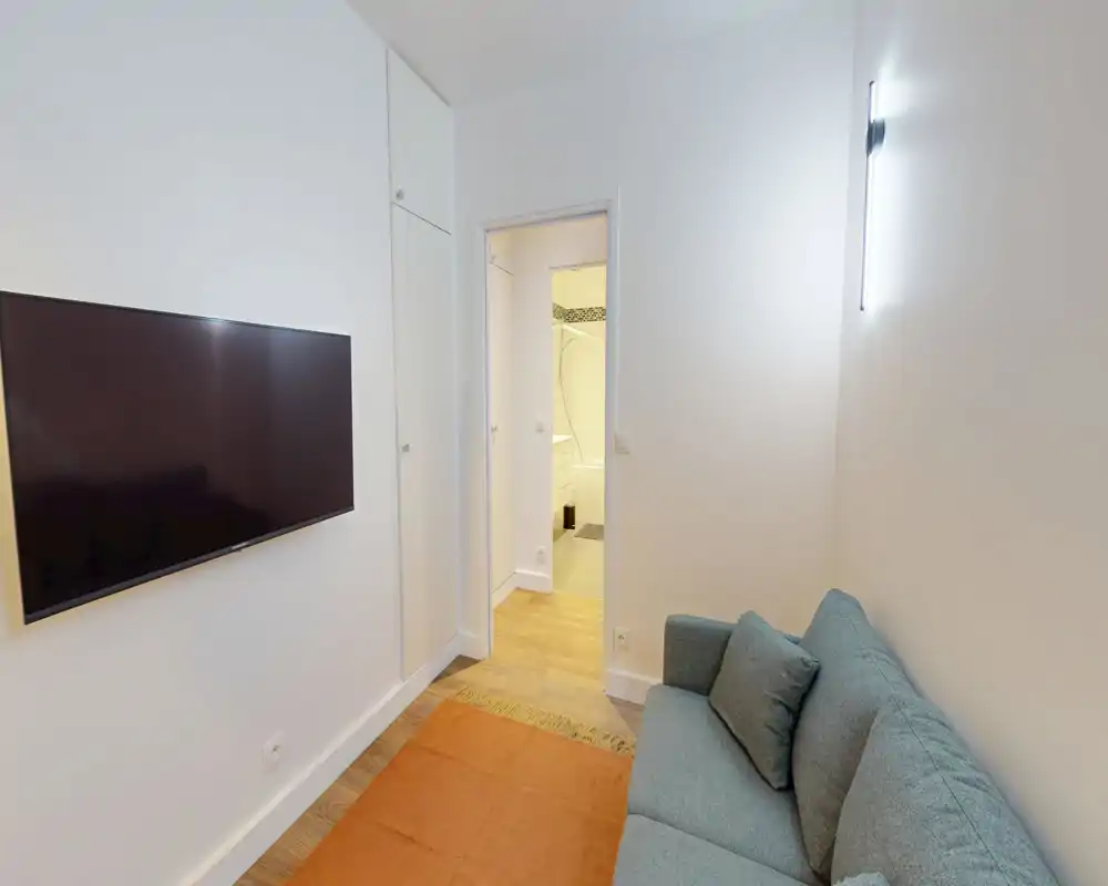 Coin salon compact avec télévision murale, canapé deux places et tapis ; murs blancs et sol en bois apportent une atmosphère sobre et confortable.