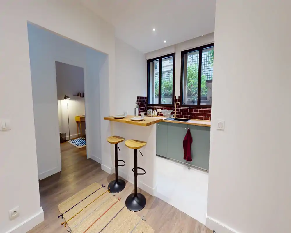 Cuisine moderne et compacte avec un bar petit-déjeuner et deux tabourets en bois, meubles bas verts, crédence en carreaux sombres et fenêtre apportant la lumière naturelle.
