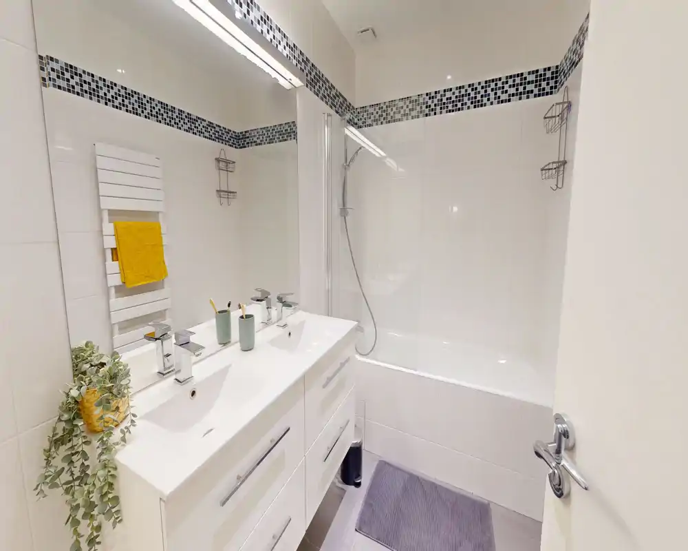 Salle de bain blanche et moderne avec baignoire-douche, meuble à double vasque, grand miroir et frise en mosaïque ; lumineux et bien rangé.