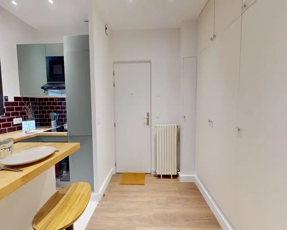 Petit hall d'entrée bien éclairé avec porte blanche, radiateur mural et rangements intégrés sur le mur droit. Le coin bar et la vue partielle sur la cuisine donnent une idée de la configuration du logement.
