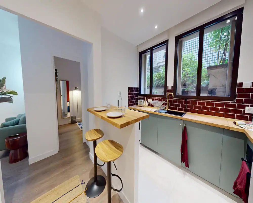 Cuisine moderne et compacte avec bar en bois, deux tabourets et placards bas verts. De larges fenêtres apportent de la lumière naturelle sur une crédence carrelée et l'évier — parfait pour un appartement urbain chaleureux.