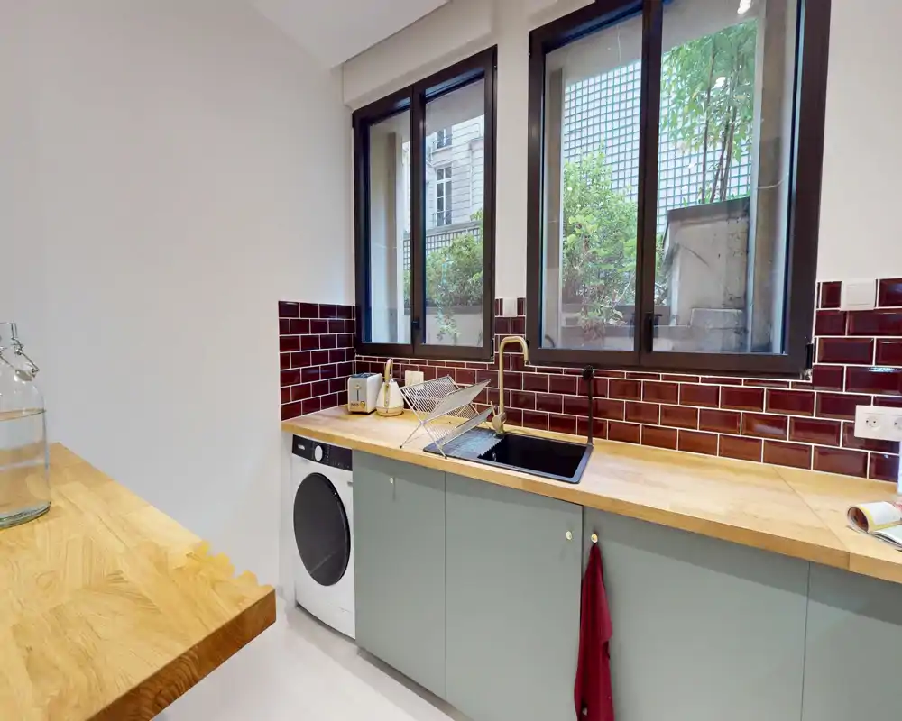 Cuisine moderne et lumineuse avec plans de travail en bois, crédence en carreaux bordeaux, évier intégré et électroménagers; grandes fenêtres apportent une lumière naturelle et une ambiance soignée.