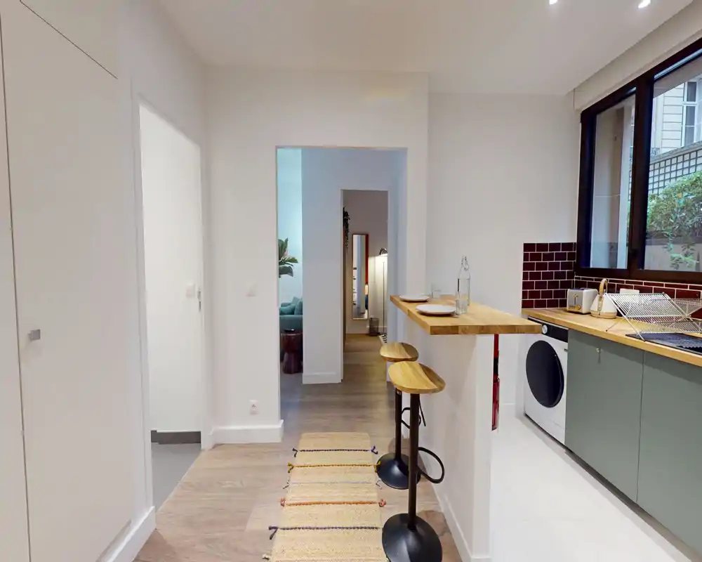 Kitchenette lumineuse et moderne avec bar en bois, deux tabourets, éléments bas verts et lave-linge sous le plan de travail — parfaite pour un appartement compact.