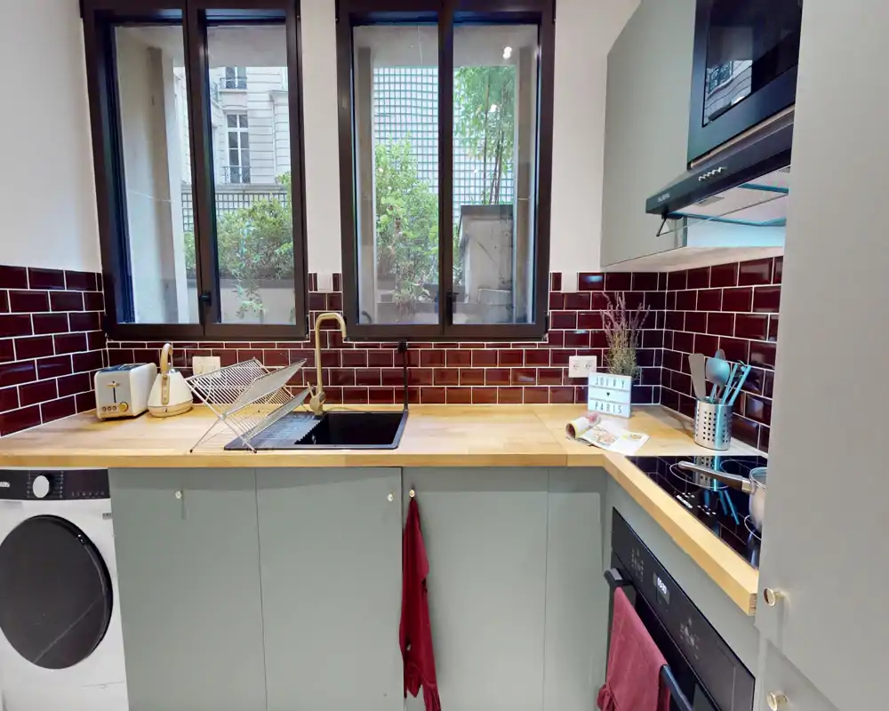 Cuisine compacte et moderne avec plan de travail bois, crédence en carreaux bordeaux et une grande fenêtre apportant de la lumière naturelle. Équipée d'un évier, plaque, four et petits appareils, fonctionnelle et élégante.