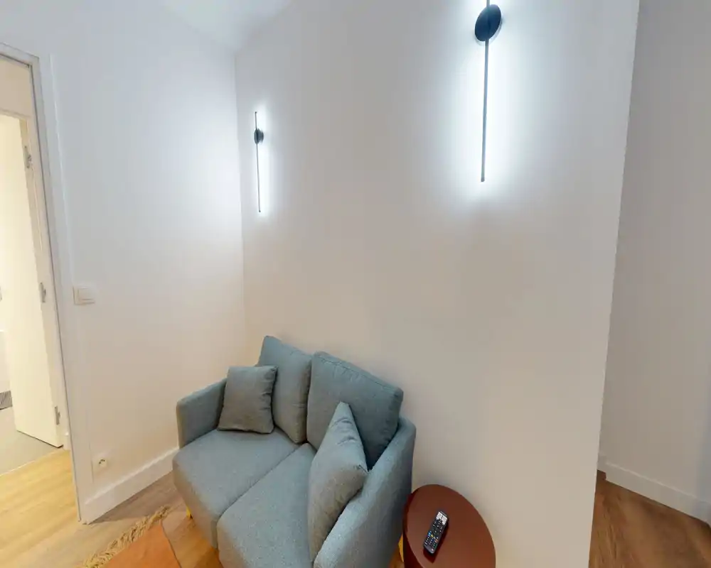 Espace salon compact avec un canapé deux places, coussins moelleux et appliques murales modernes ; tons neutres et sol en bois pour une ambiance cosy et minimaliste.