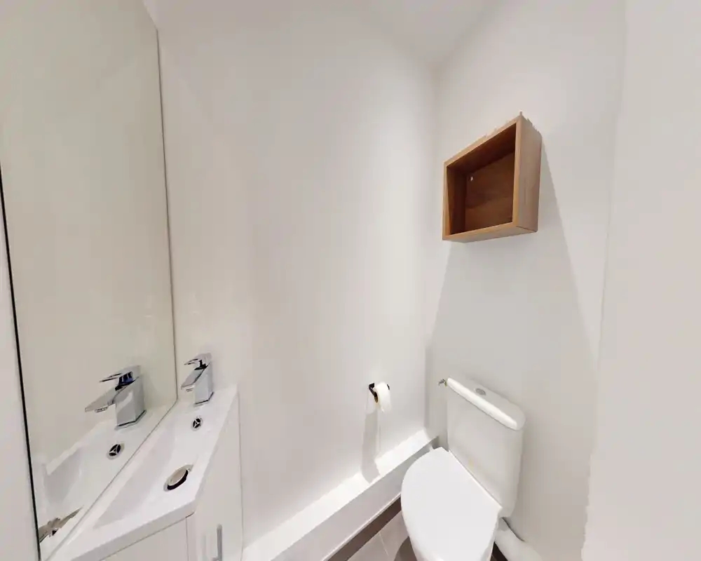 Salle de bains compacte et moderne aux murs blancs, toilettes et lavabo d'angle avec miroir ; petite étagère en bois minimaliste.