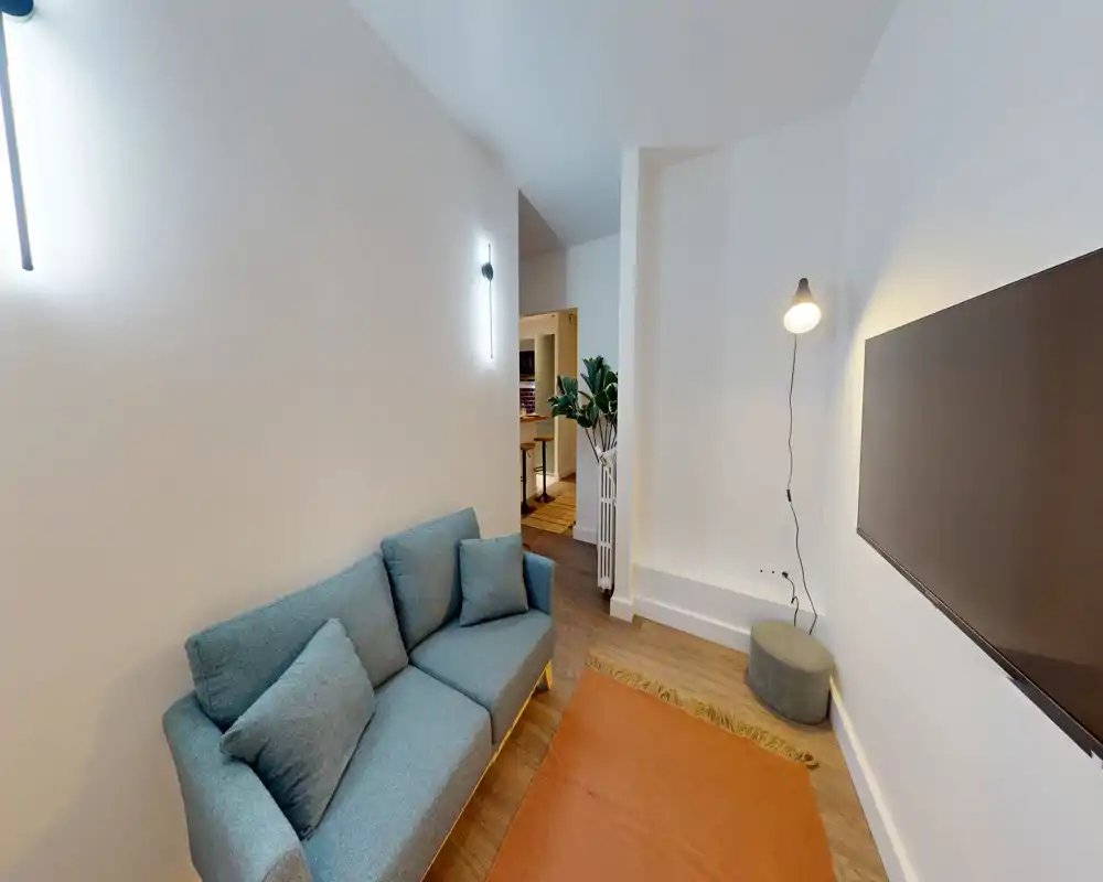 Salon moderne et chaleureux avec un canapé bleu, télévision murale et éclairage doux ; parquet et petit tapis créent un coin détente accueillant.