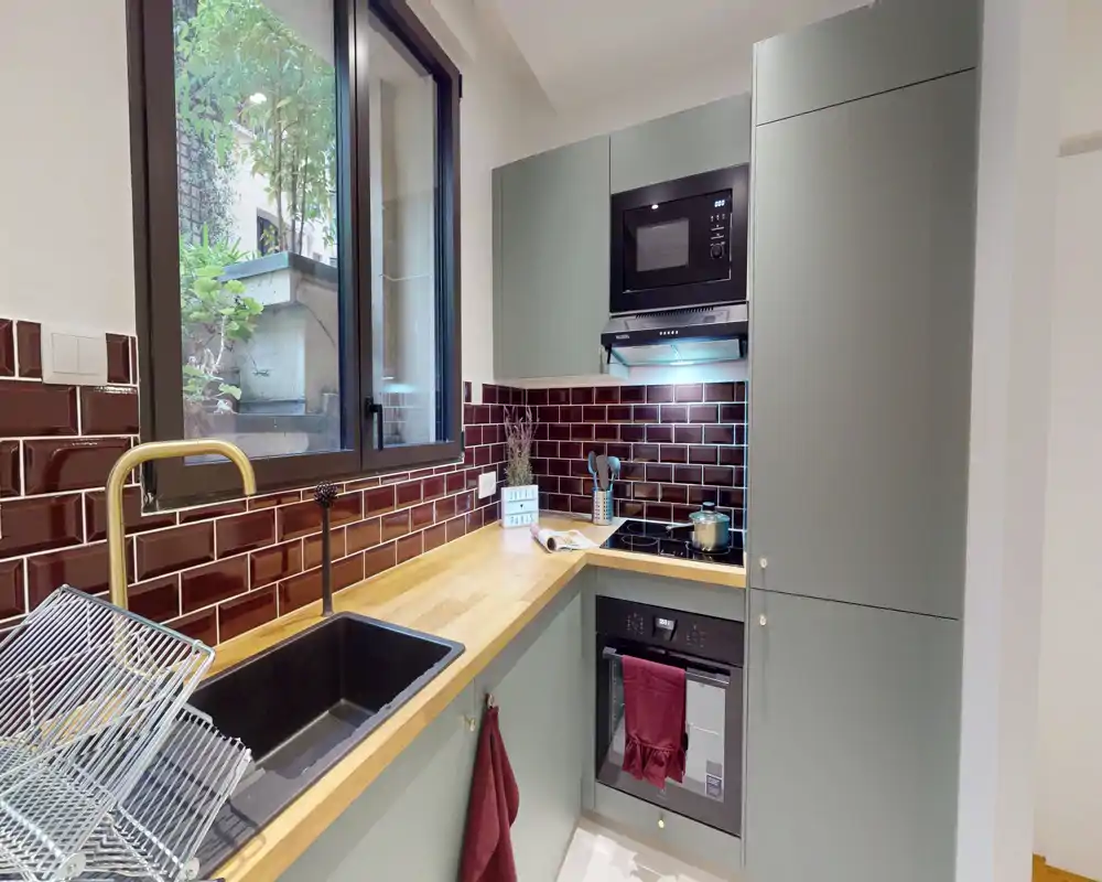 Cuisine moderne et compacte avec plan de travail en bois, crédence en carreaux bordeaux, four et micro-ondes encastrés, et une grande fenêtre apportant la lumière naturelle.