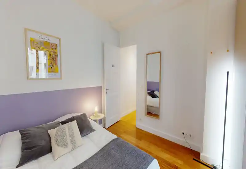 Chambre lumineuse et décorée avec lit double, coussins décoratifs, lampe de chevet et parquet en bois ; mur accent lilas et miroir en pied pour une ambiance moderne et chaleureuse.