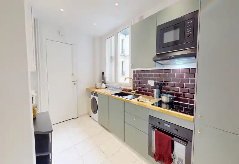 Cuisine en long moderne avec placards verts, plan de travail en bois, four et micro-ondes encastrés et évier sous une fenêtre lumineuse — un espace compact et élégant, parfait pour cuisiner efficacement.