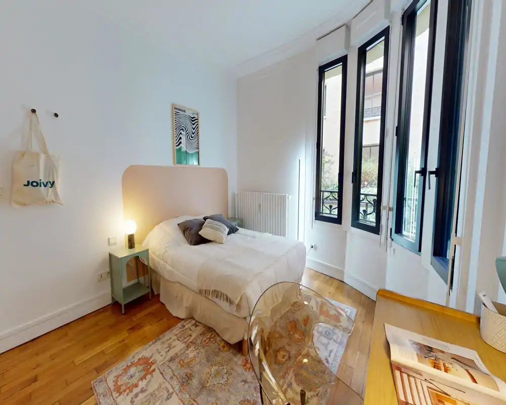 Chambre lumineuse et cosy avec lit double, table de chevet et grandes fenêtres apportant une belle lumière naturelle; tapis et textiles doux pour une ambiance chaleureuse.
