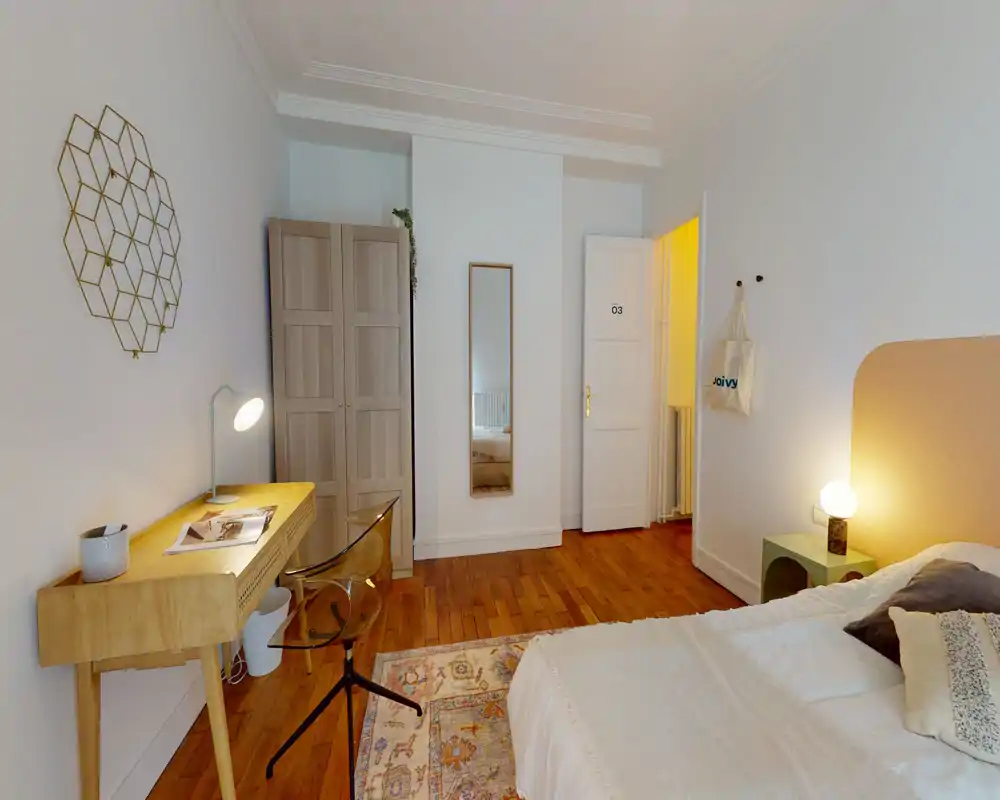 Chambre lumineuse et accueillante avec un lit confortable, parquet, bureau et armoire. L'éclairage chaud et le tapis décoratif offrent une atmosphère agréable.
