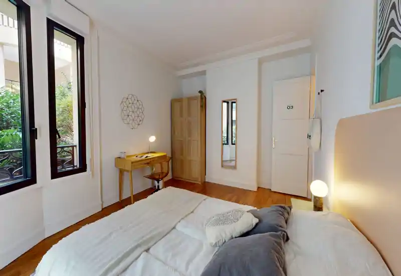 Bright, well-styled bedroom with a large bed in the foreground, warm bedside lighting and a small workspace by the window. Hardwood floors and soft textiles create a cozy, inviting atmosphere.