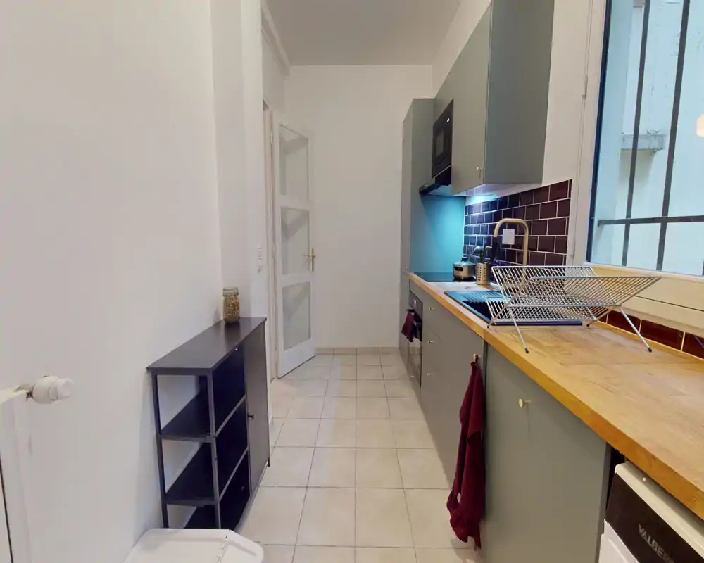 Cuisine lumineuse et étroite avec plan de travail en bois, placards gris et crédence carrelée. L'espace est propre et bien éclairé par une fenêtre latérale, avec rangements et appareils sur un seul mur.