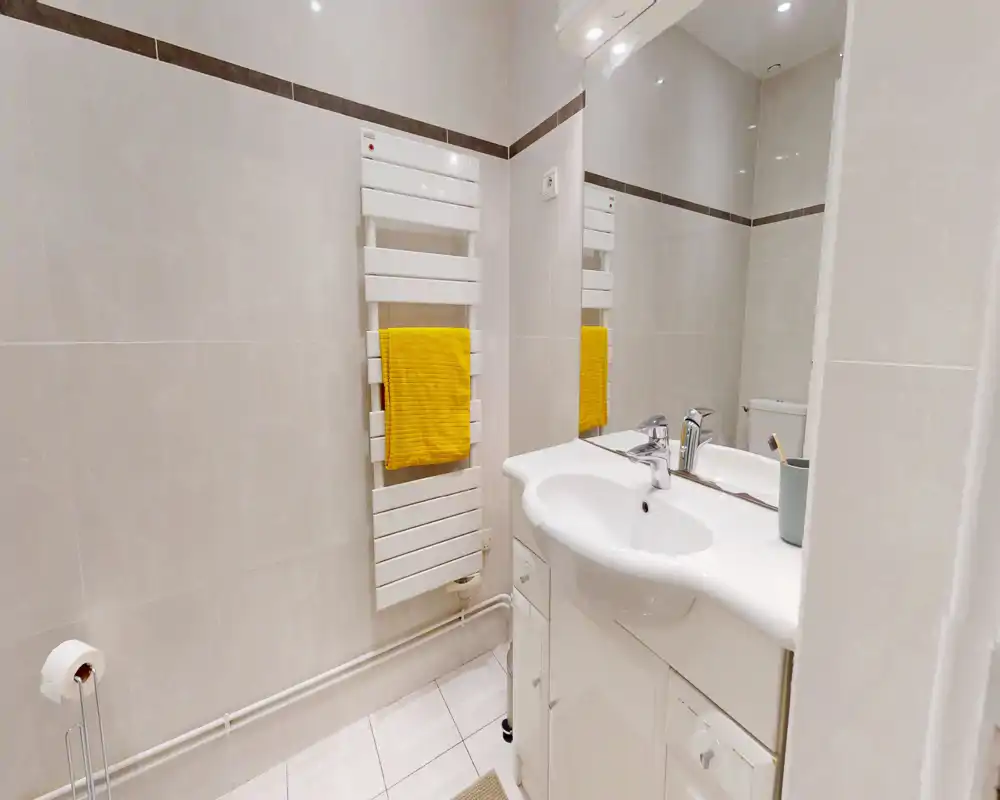 Salle de bains compacte et moderne avec carrelage blanc, meuble lavabo, radiateur porte-serviette mural avec une serviette jaune et grand miroir.