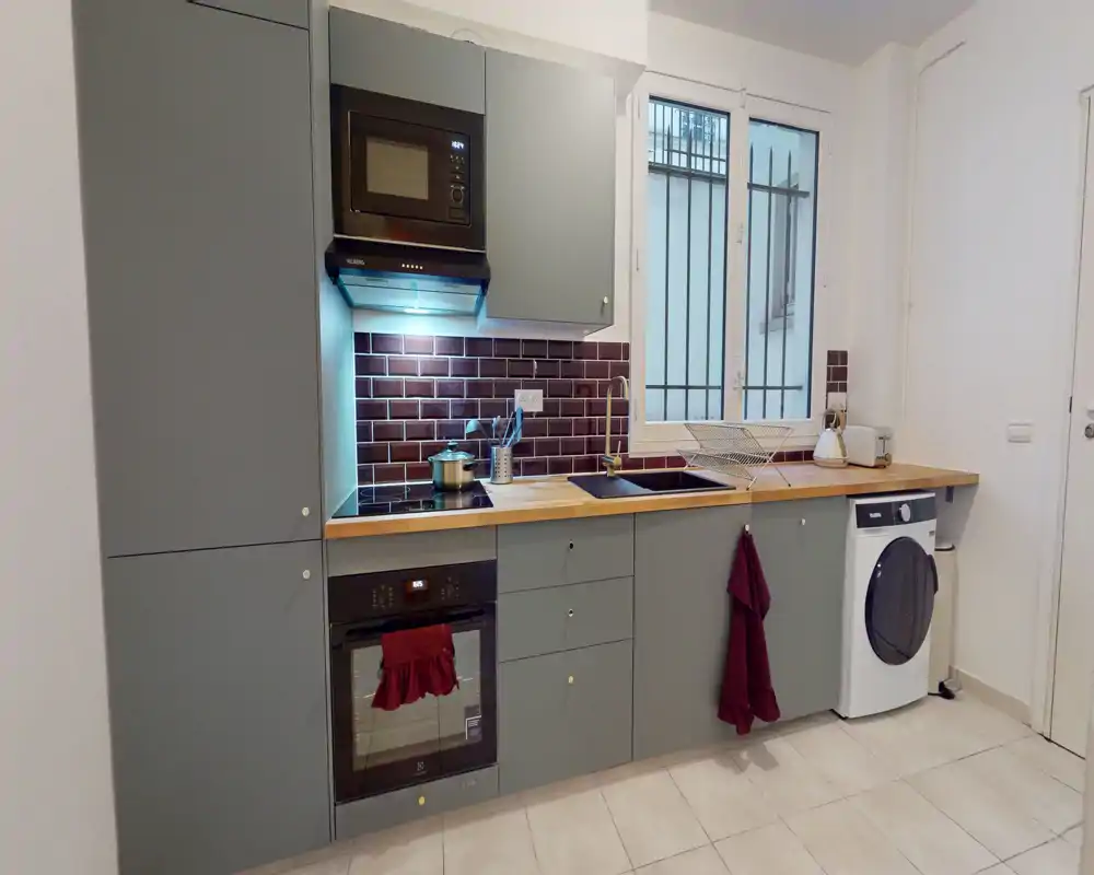 Cuisine compacte moderne avec placards gris, plan de travail en bois, four et micro-ondes encastrés, évier sous la fenêtre et lave-linge intégré — lumineuse et bien rangée.