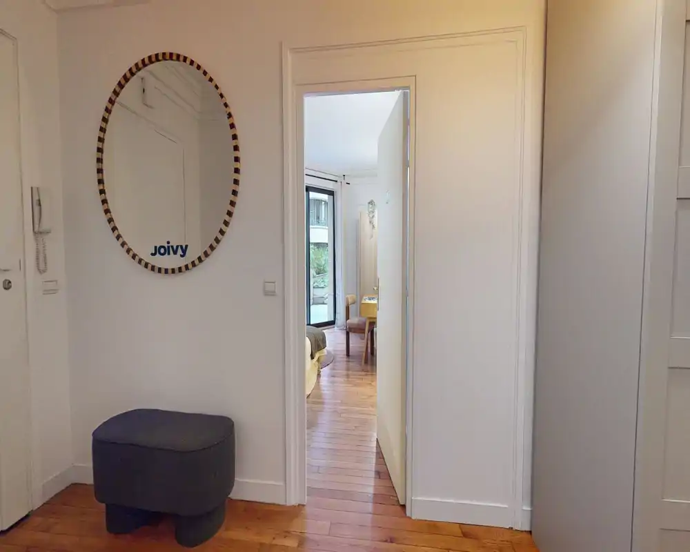 Entrée accueillante avec parquet, un miroir rond décoratif et un petit pouf capitonné ; la porte s'ouvre sur une pièce adjacente lumineuse.