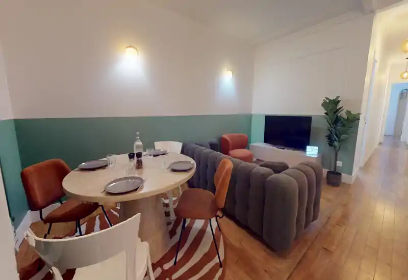 Stylish living area with a curved sofa, TV and a round dining table — warm wood floors and green-accented walls create a cozy, modern atmosphere.