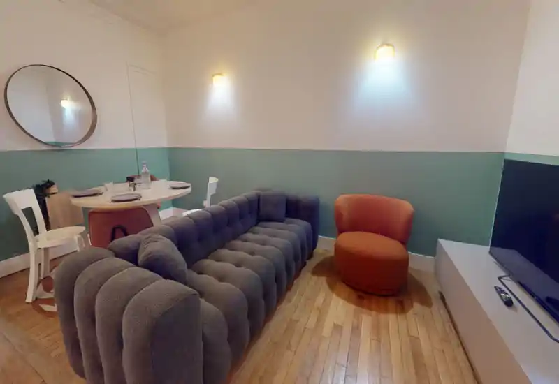 Stylish living room with a plush tufted grey sofa, orange accent chair, wall sconces and a small dining nook — warm, modern and inviting.