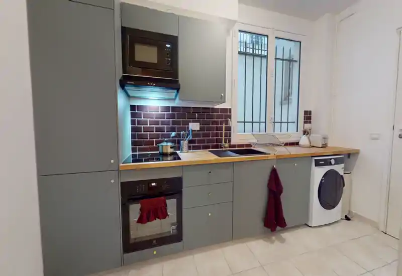 Modern compact kitchen with grey cabinets, wooden worktop, built-in oven and microwave, sink under a window and an integrated washing machine — bright and neatly arranged.