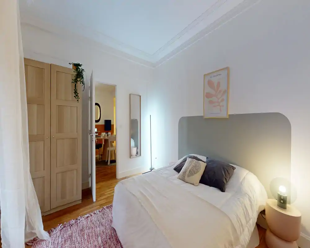 Chambre lumineuse et élégante avec un lit double confortable, textiles doux et parquet chaleureux. Elle comprend une tête de lit arrondie, une table de chevet avec lampe, une armoire et des éléments décoratifs accueillants.