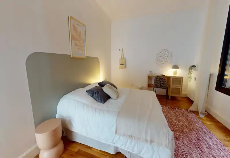 Chambre lumineuse et chaleureuse avec lit double bien fait, coussins décoratifs, tapis texturé et un petit bureau dans le coin — idéale pour un séjour confortable.
