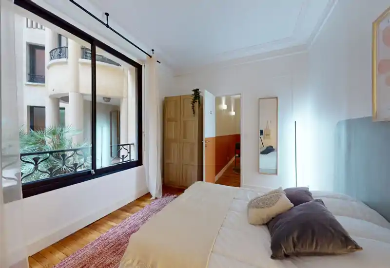 Chambre lumineuse et soignée avec grande fenêtre sur cour intérieure, parquet, lit double confortable avec coussins superposés et tapis texturé.