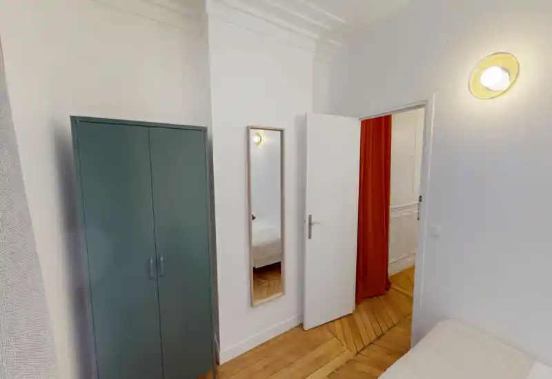 Coin de chambre compact avec une armoire fine, un miroir en pied et un aperçu du lit par la porte ouverte. Murs blancs, parquet chaleureux et éclairage mural doux pour une ambiance calme et minimaliste.