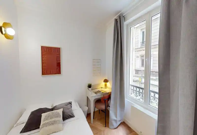 Chambre simple et lumineuse, lit bien fait, lampe de chevet et petit bureau près d'une grande fenêtre avec vue sur la rue — parfaite pour un voyageur seul.