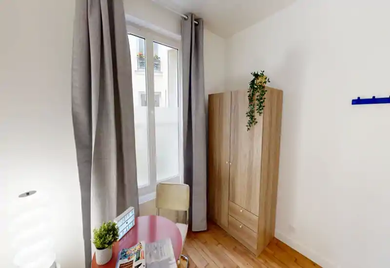 Coin chambre avec armoire, bureau et grande fenêtre habillée de rideaux. Lumière naturelle abondante et mobilier simple et moderne pour un espace soigné et fonctionnel.