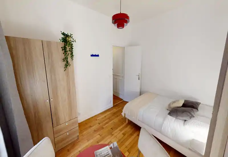 Chambre simple lumineuse et bien aménagée avec murs blancs, parquet en bois et lit confortable en linge neutre ; armoire et petit espace assis rendent la pièce chaleureuse.