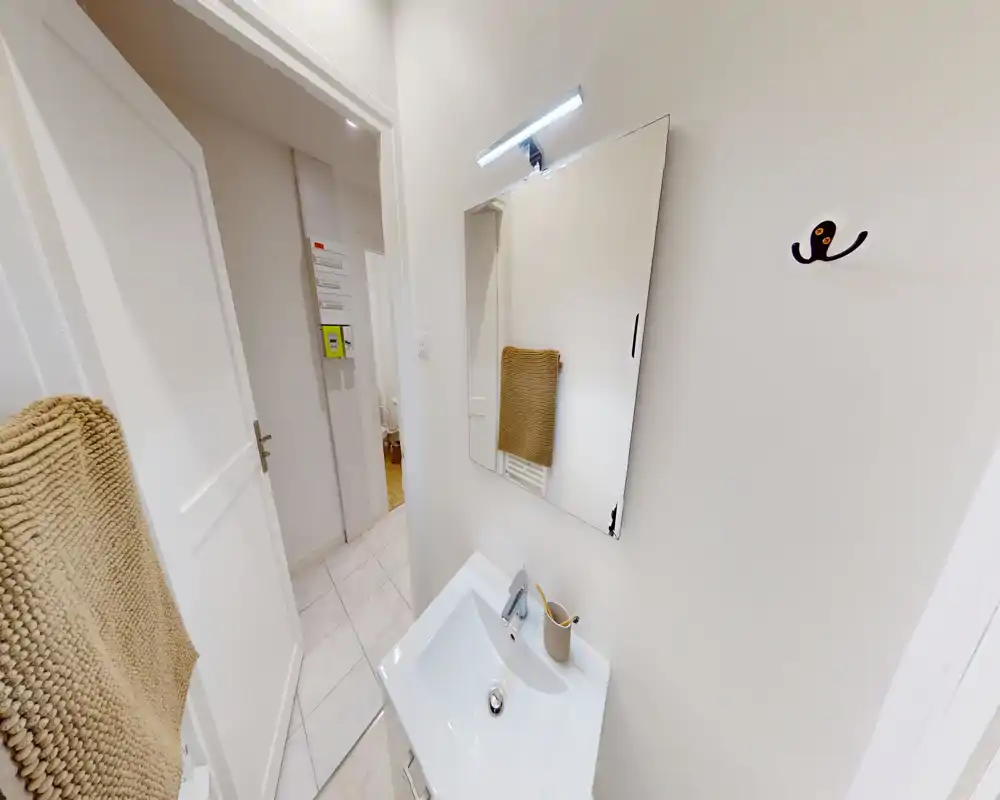 Angolo bagno compatto e ben illuminato con lavabo bianco, specchio e asciugamano; finiture moderne e tonalità neutre.