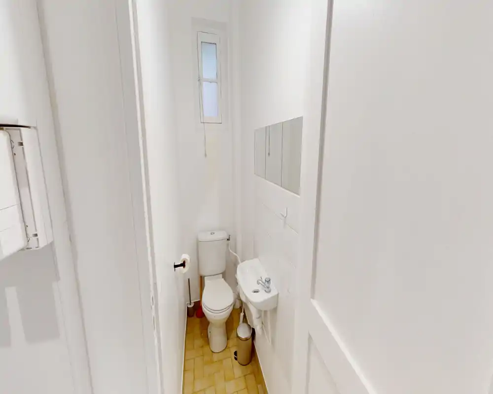 Bagno compatto e ben illuminato con WC, piccolo lavabo a muro e una finestra stretta con vetro satinato. Pareti bianche e pavimento chiaro rendono lo spazio luminoso e funzionale, ideale come bagno di servizio.