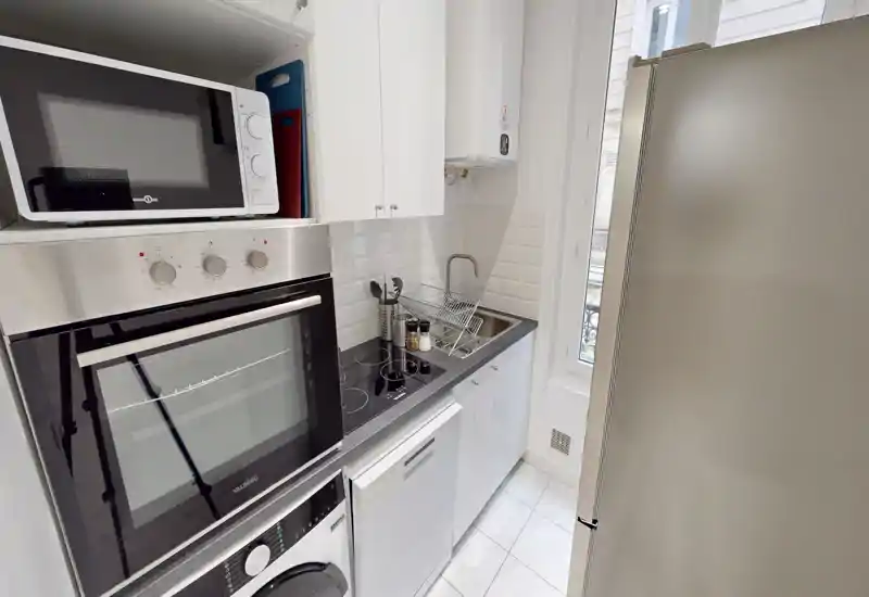 Cuisine moderne et compacte avec four encastré, micro-ondes, plaque à induction, évier et grand réfrigérateur ; placards blancs et crédence carrelée pour une belle luminosité.