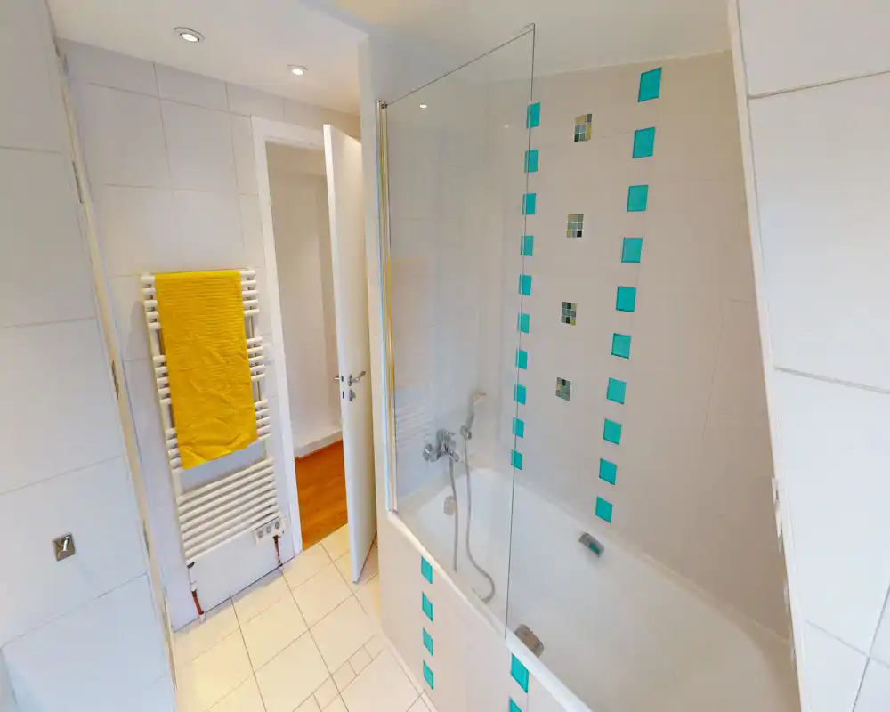 Modern, bright bathroom with a bathtub-shower combo, glass screen and decorative turquoise tiles. White wall tiles and a heated towel rail with a yellow towel make the space fresh and inviting.