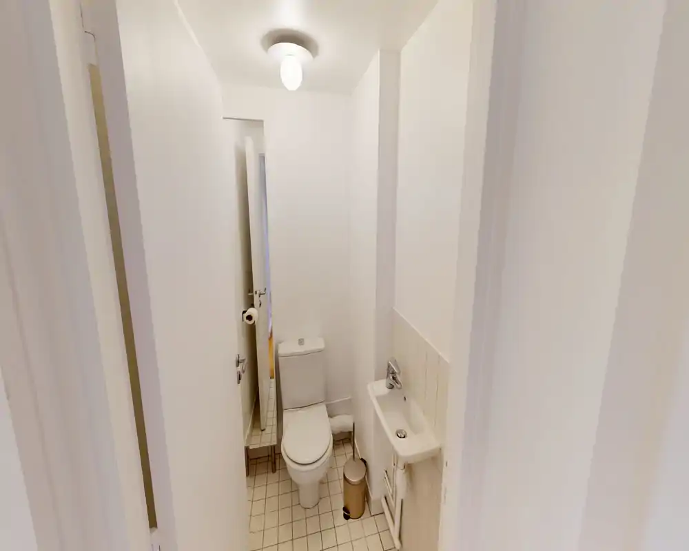 Compact bathroom with a close-coupled toilet, small wall-mounted sink, tiled floor and a mirror beside the door. Bright, neutral decor suitable for a guest or en-suite washroom.