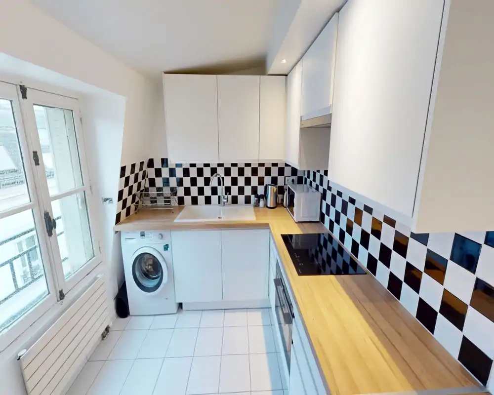 Bright modern kitchen with wood countertops, black-and-white checkerboard backsplash, integrated stovetop and a washing machine under the counter — clean, functional layout ideal for compact apartments.
