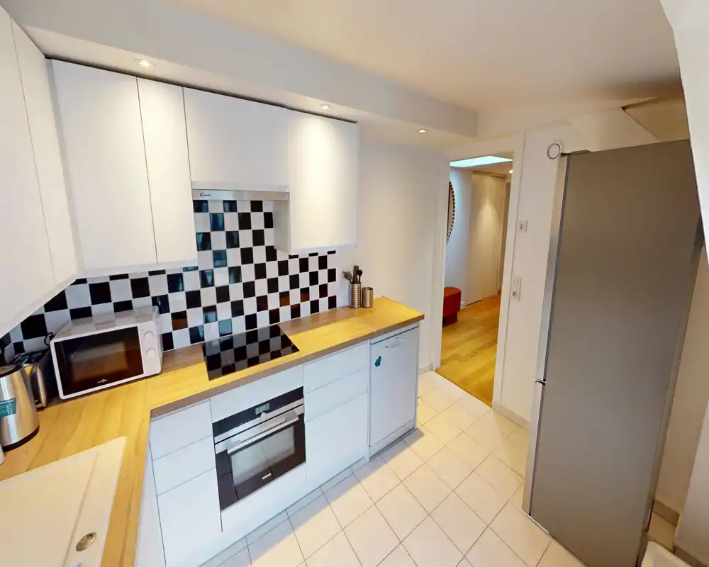Modern compact kitchen with white cabinets, wood-effect countertops and a black-and-white tiled splashback. Equipped with an oven, induction hob, microwave and tall fridge; bright, tidy and efficiently arranged for city apartments.