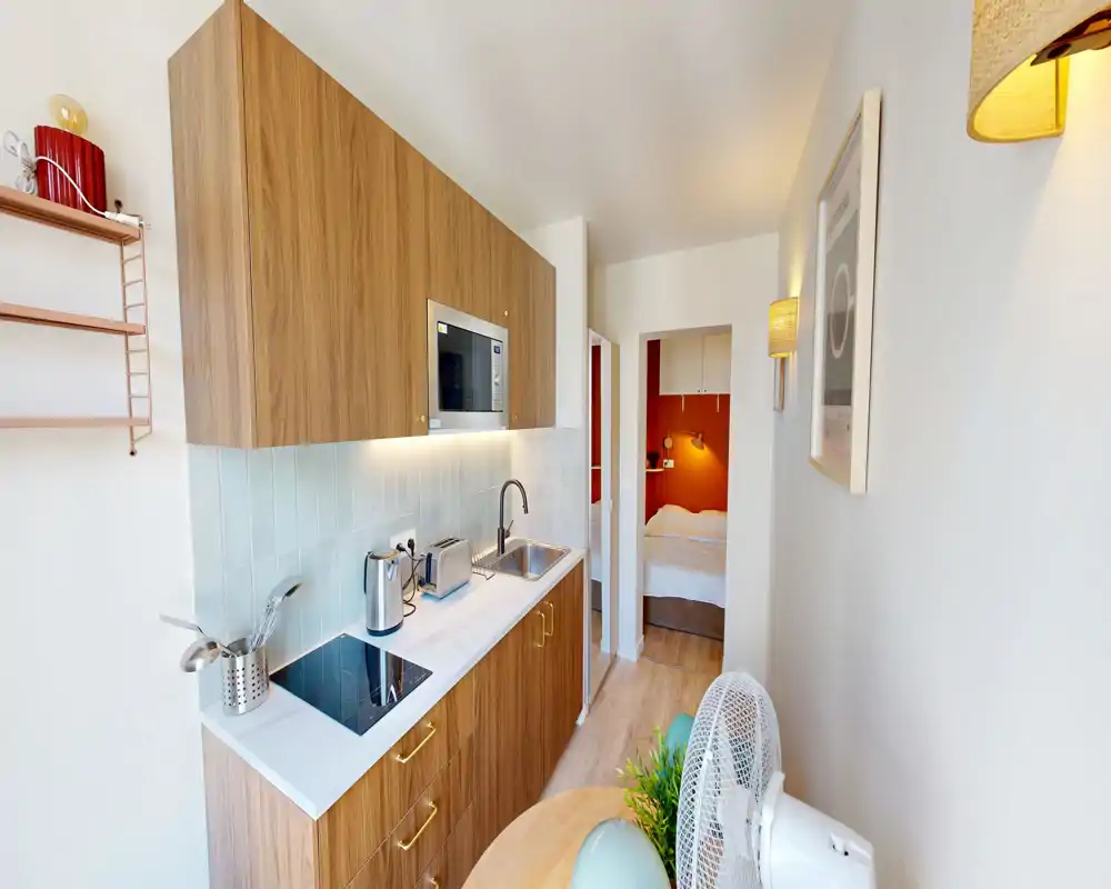 Kitchenette compacte aménagée avec armoires en bois, plaque de cuisson et micro-ondes intégrés, petit coin repas et lumière chaude et lumineuse — idéale pour un studio.