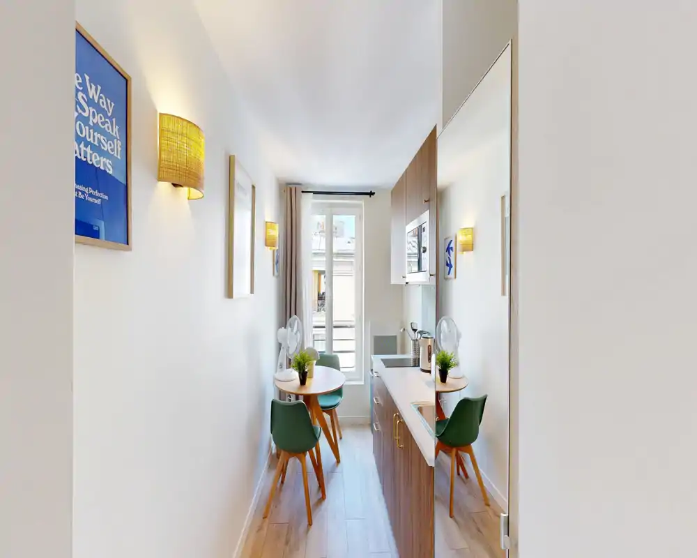 Cuisine moderne compacte à disposition étroite : placards en bois, plan de travail blanc, petite table avec chaises vertes et fenêtre lumineuse apportant la lumière naturelle.