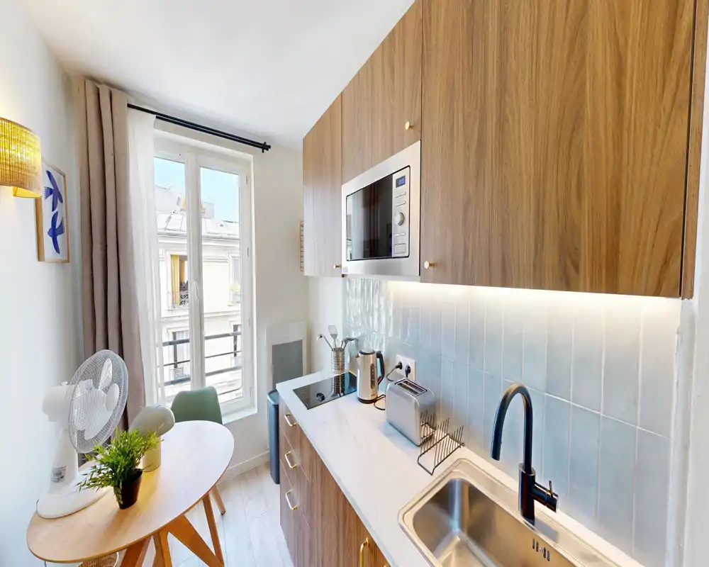 Cuisine compacte et moderne avec placards en bois, plan de travail blanc, micro-ondes encastré, plaque à induction et évier ; une petite table ronde près de la fenêtre offre un coin petit-déjeuner et lumière naturelle.