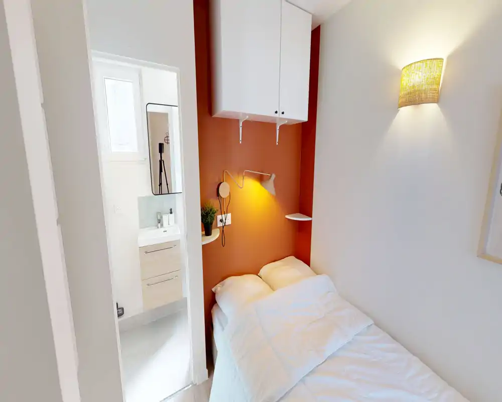 Chambre compacte avec un lit simple près d'un mur d'accent terracotta, lampe de chevet et étagères murales ; pièce claire et fonctionnelle avec une salle de bains attenante visible.