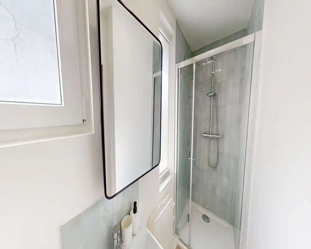 Salle de bains compacte et moderne avec douche vitrée, miroir armoire mural et petit lavabo ; carreaux gris clair et appareils sanitaires blancs pour un rendu propre et lumineux.